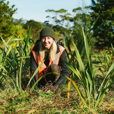 Woman in beanie and hi vis crouching amongst Harakeke plants