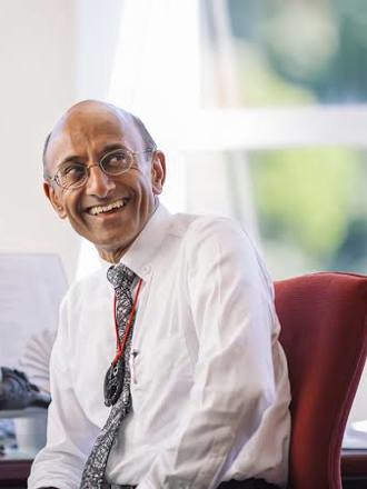 Smiling man in white button up with black and white tie turned in seat with desk with papers and Maori artifacts on it.